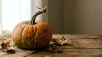 Orange speckled pumpkin with curved stem on rustic wooden table beside autumn leaves creates warm seasonal harvest decoration for fall celebrations.