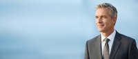 Confident mature businessman with gray hair in dark suit smiling outdoors against blue sky background for corporate leadership and professional success concepts.
