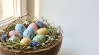 Colorful Easter eggs in wicker basket with dried grass and blue flowers on white table near window. Spring holiday decoration for greeting cards.