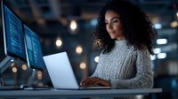Young African American woman software developer working late at office typing on laptop computer with code on multiple monitors in modern workspace.
