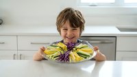 Happy young boy with tousled brown hair smiling while holding colorful plate of fresh fruit slices in bright modern kitchen for healthy eating concepts.