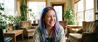 Happy young Asian woman with colorful hair laughing in cozy living room with plants and natural light for lifestyle and wellness content.