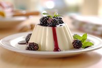 Creamy vanilla panna cotta topped with fresh blackberries and mint leaves, drizzled with berry sauce on white plate, selective focus with soft background.