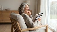 Senior woman with gray hair relaxing in wooden chair using digital tablet for video call at home. Modern living room interior with natural light.