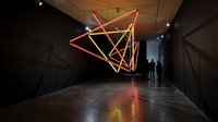 Modern art gallery interior with geometric neon light installation creating abstract triangular shapes in red and yellow colors on dark walls.
