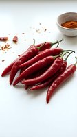 Fresh red chili peppers arranged on white surface with ground spice powder in ceramic bowl, ideal for cooking, food styling, and culinary content.