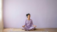 Woman in lavender shirt meditating cross-legged on yoga mat against purple wall, practicing mindfulness and wellness for relaxation and fitness.