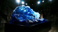 Large blue translucent wave sculpture displayed in dark museum gallery with dramatic spotlighting creating artistic shadows and reflections on walls.