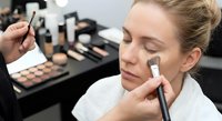 Professional makeup artist applying foundation to young Caucasian woman client in beauty salon with cosmetic products and brushes on table.