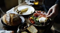 Mediterranean appetizer platter with fresh bread, prosciutto, cheese, olives and cherry tomatoes served on rustic wooden table in moody lighting for food photography.