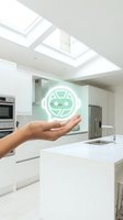 Hand holding glowing green AI robot face hologram in modern white kitchen with skylights showcasing smart home automation technology and artificial intelligence.