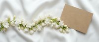 Blank kraft paper card with white flower garland on soft fabric background, suitable for wedding invitations, greeting cards, and event announcements.