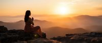 Woman silhouette meditating on mountain peak during golden sunset with layered hills in background creating peaceful spiritual moment for wellness and mindfulness.
