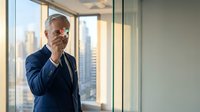 Senior businessman in blue suit talking on smartphone in modern office with city skyline view through large windows for corporate communication.
