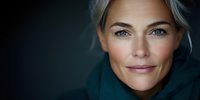 Confident mature woman with natural gray hair and bright eyes wearing teal sweater smiling against dark background in dramatic lighting portrait.