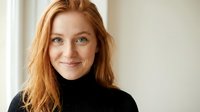 Young redhead woman with freckles wearing black turtleneck sweater smiling at camera in bright indoor setting with natural lighting for lifestyle content.