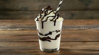 Creamy vanilla milkshake topped with whipped cream and chocolate syrup drizzle in glass with striped straw on rustic wooden table background.