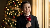 Young Caucasian boy in formal black suit with red bow tie smiling at camera during Christmas celebration with decorated tree and warm lights in background.
