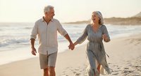 Happy senior couple walking hand in hand on beach during golden hour sunset, enjoying romantic vacation together in casual summer clothing.