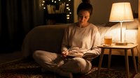 Young woman relaxing at home in evening, sitting on couch with smartphone and warm tea, cozy living room with soft lamp lighting creating peaceful atmosphere.
