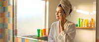 Middle-aged woman with light skin tone and blonde hair wrapped in towel, wearing white bathrobe, applying skincare product to face in modern bathroom with colorful cosmetic bottles on shelves and counter.