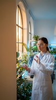 Young female scientist in white lab coat examining DNA structure hologram in modern laboratory with plants and natural lighting for biotechnology research.