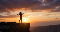 Silhouette of woman celebrating success on mountain cliff edge at golden sunset with dramatic orange sky and valley landscape below.