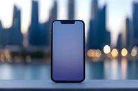 Modern smartphone with blank screen against blurred urban cityscape background with bokeh lights and water reflection during blue hour.