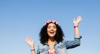 Young woman with long curly dark hair and medium skin tone wearing denim jacket and flower crown, arms raised in celebration against clear blue sky.