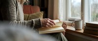 Elderly woman reading a book by a window in a cozy living room, with a warm sweater, coffee mug, and stack of books nearby.