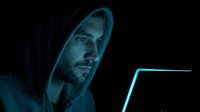 Man in hoodie with neon blue lighting working on laptop against dark background. Cybersecurity, hacking, technology crime concept for digital security and online threat awareness.