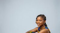 Young woman with medium skin tone, braided hairstyle, wearing blue beaded necklace, smiling at camera against neutral gray background for portrait and lifestyle use.