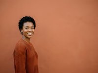 Young African American woman with short curly hair wearing a rust-colored ribbed sweater, smiling at camera against warm terracotta background. Portrait for lifestyle, fashion, and diversity marketing.