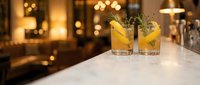 Two golden cocktails with rosemary garnish on marble bar counter in upscale restaurant with warm ambient lighting for hospitality and dining concepts.