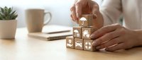 Hands of a woman stacking wooden cubes with insurance icons including home, car, life and health symbols on a desk.