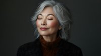 Elegant mature Asian woman with silver hair and closed eyes wearing brown turtleneck sweater against dark background, expressing serenity and confidence.