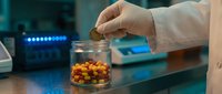 Scientist in white coat examining colorful pharmaceutical pills and capsules in glass container during laboratory research and drug development process.