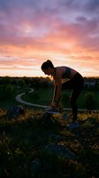 Female athlete in athletic wear stretching outdoors at sunset, silhouetted against golden sky with suburban landscape below, ideal for fitness and wellness content.