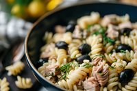 Fusilli pasta salad with tuna chunks, black olives and fresh herbs in dark bowl. Close up view of Mediterranean style cold pasta dish with selective focus.
