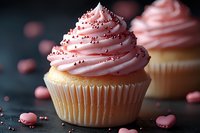 Vanilla cupcake topped with swirled pink frosting and silver dragees, decorated with scattered heart-shaped candies on dark background. Food photography for bakery marketing.