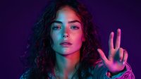 Young woman with curly hair in neon lighting showing peace sign gesture against dark background, dramatic red and blue color contrast creates modern cyberpunk atmosphere.