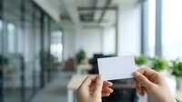 Hands holding blank white business card in modern office corridor with glass partitions and green plants, suitable for corporate branding and professional networking concepts.