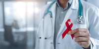 Healthcare professional in white coat holding red AIDS awareness ribbon in modern medical facility with stethoscope around neck.