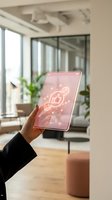 Hand holding pink tablet device with glowing neon planet and rocket icon display in modern office workspace with windows, plants, and contemporary furniture for technology and innovation concepts.