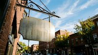 Blank vintage metal sign hanging from ornate iron bracket on brick building wall in historic downtown district with blue sky and urban street scene.