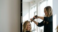 Professional female hairstylist curling blonde woman's hair with styling iron in bright modern salon interior with natural window lighting.