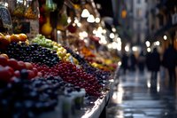 Fresh colorful berries and fruits displayed at outdoor market stall with blurred bokeh city lights and people walking in background, selective focus on produce.