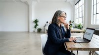 Mature businesswoman with glasses working on laptop in modern office space with large windows and minimalist design for corporate presentations.