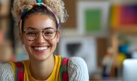 Young mixed race woman with curly blonde hair and glasses smiling warmly at camera, wearing colorful casual attire in home office setting. Natural lighting highlights genuine expression.