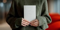 Woman in green knitted sweater holding blank white book or journal with manicured nails against blurred background.